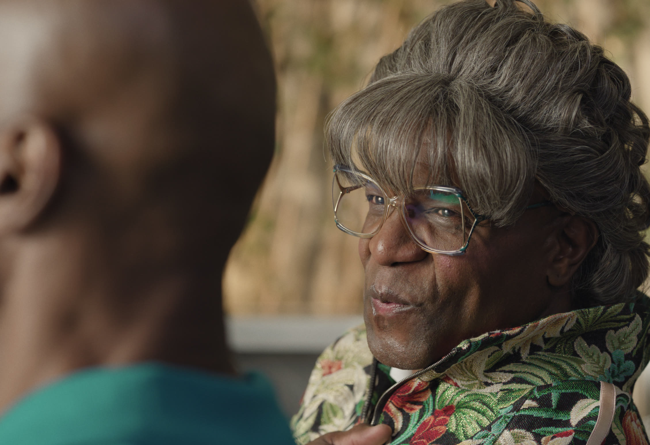 Two people in conversation, one wearing a grey wig, glasses, and a floral jacket
